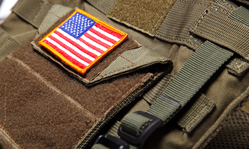 Close-up of a U.S. flag patch attached to a tactical vest made of brown and green fabric with reinforced stitching and straps.