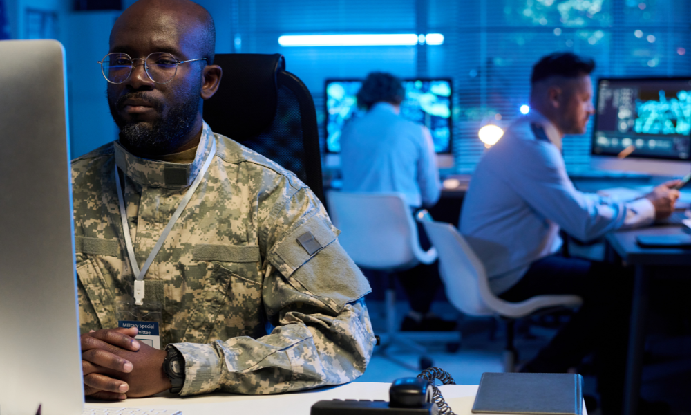 Soldier in digital camouflage uniform working on a desktop computer in a high-tech operations room with multiple monitors and blue lighting.