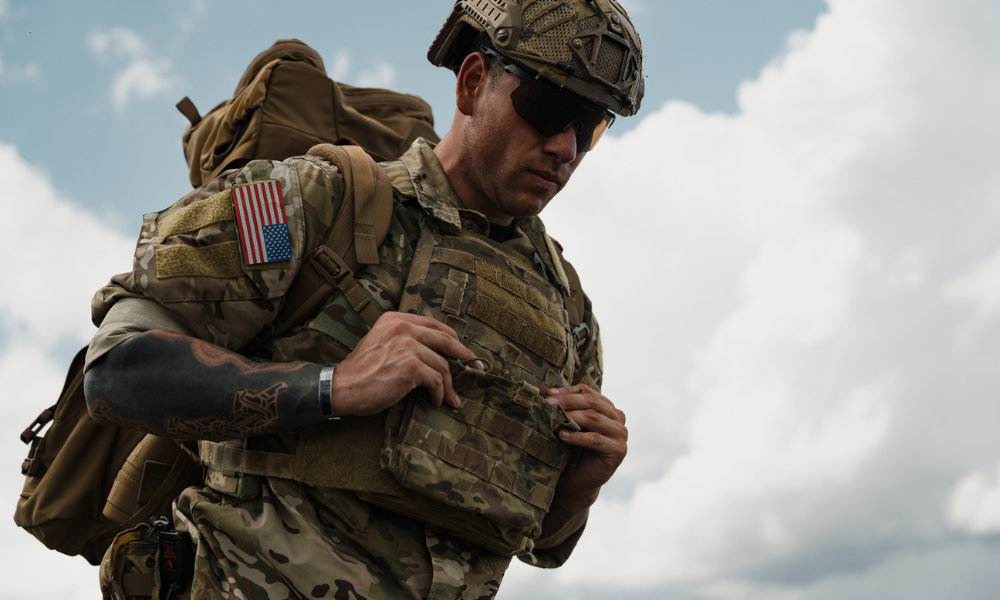 Heavily equipped American soldier adjusting his tactical vest, wearing a helmet and sunglasses under cloudy sky.