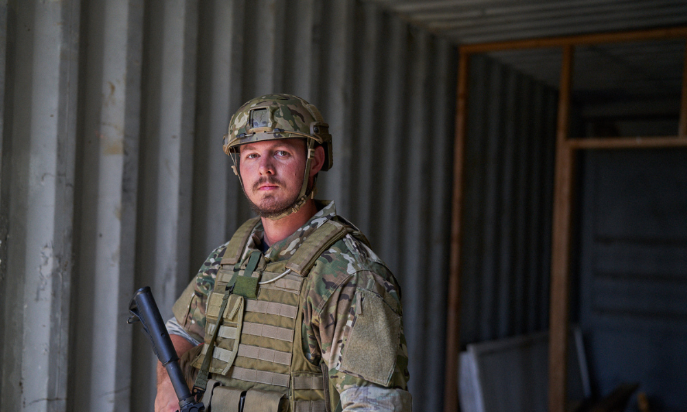 Male soldier in full combat gear standing indoors, holding a rifle, and looking ahead with serious expression.