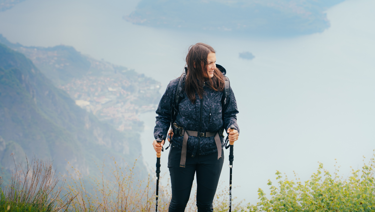 Donna con zaino e bastoncini da trekking su un sentiero in quota, con lago di garda sullo sfondo.