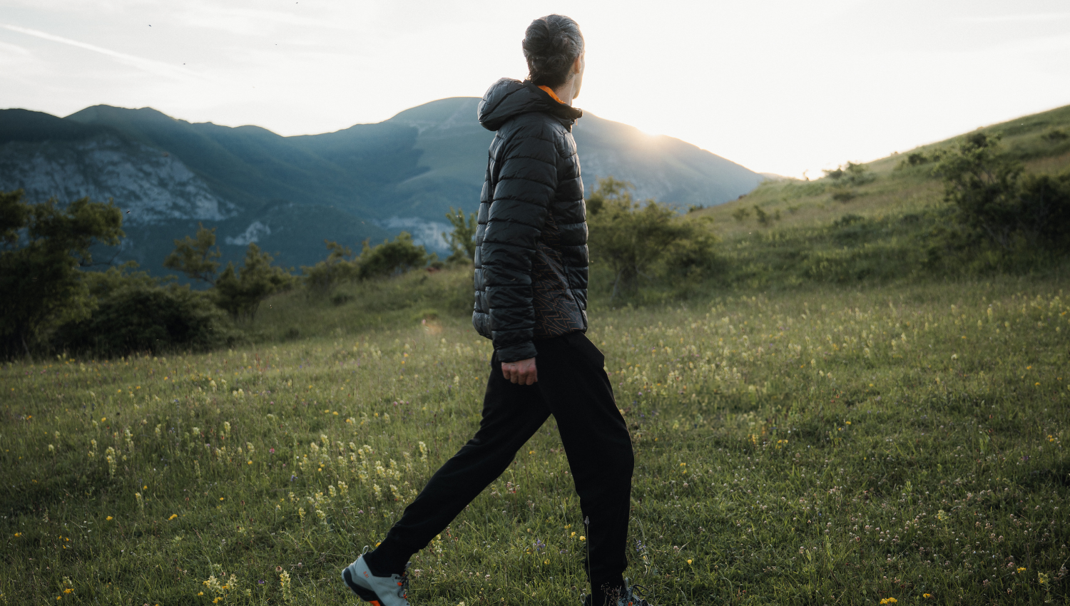 Persona che cammina su un prato di montagna al tramonto, con colline e montagne sullo sfondo.