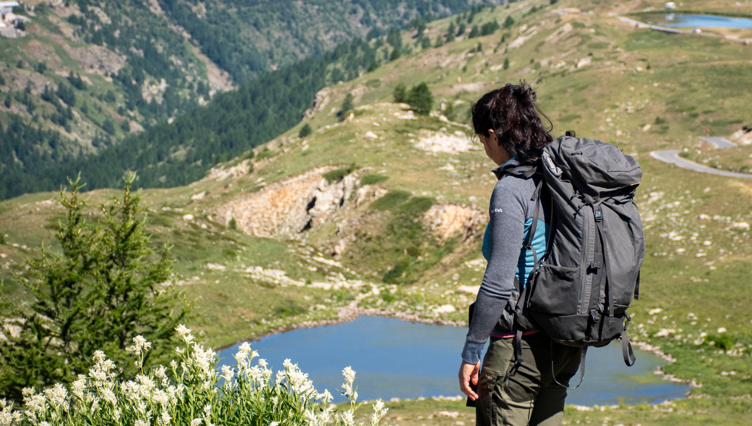 Mujer con mochila observando un lago alpino desde un sendero en la montaña, rodeada de prados y colinas.
