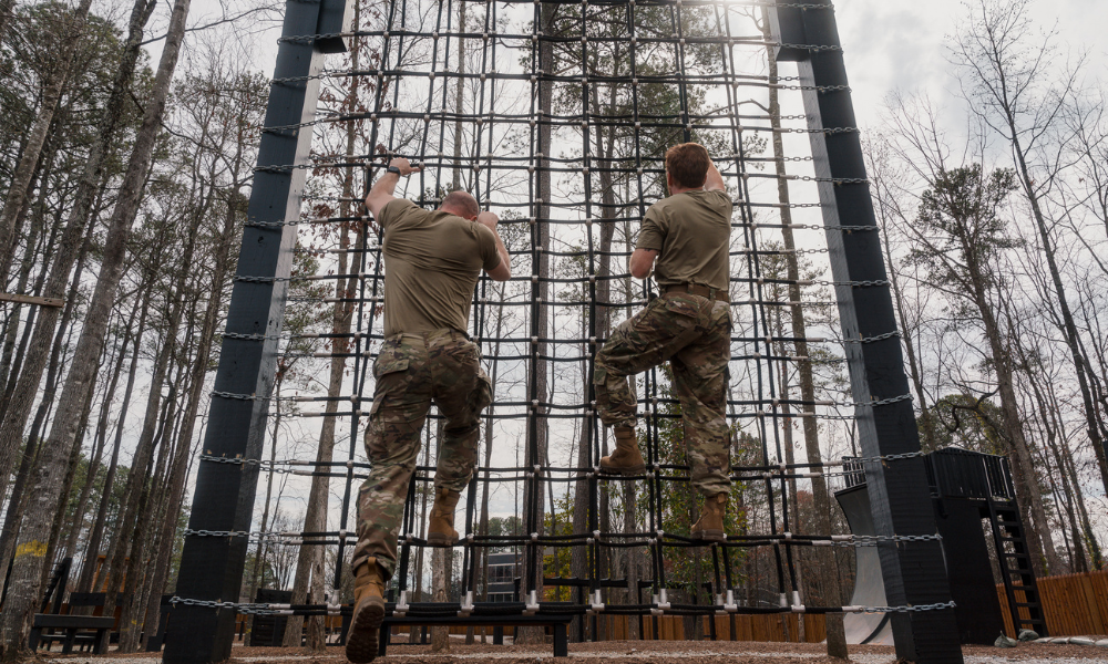 Two soldiers climbing a rope wall obstacle during military training in a wooded area.