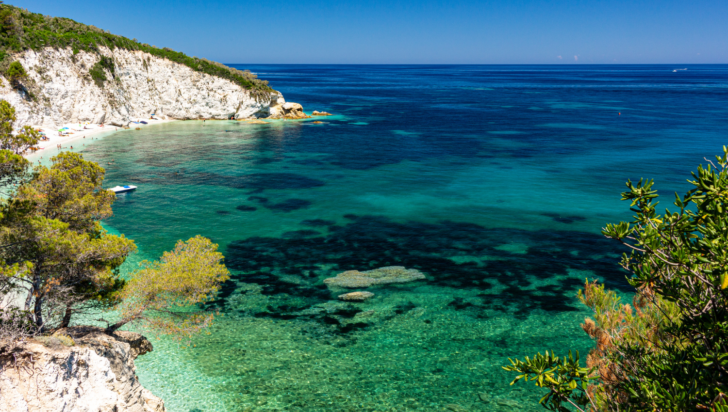 Baia isola d'elba con acqua cristallina e spiaggia affollata, circondata da scogliere verdi e mare turchese.