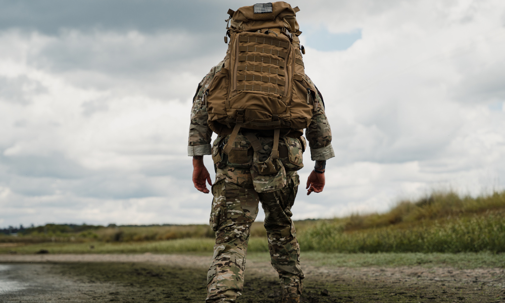     Rear view of a soldier carrying a large tactical backpack while walking across rugged terrain under cloudy skies.