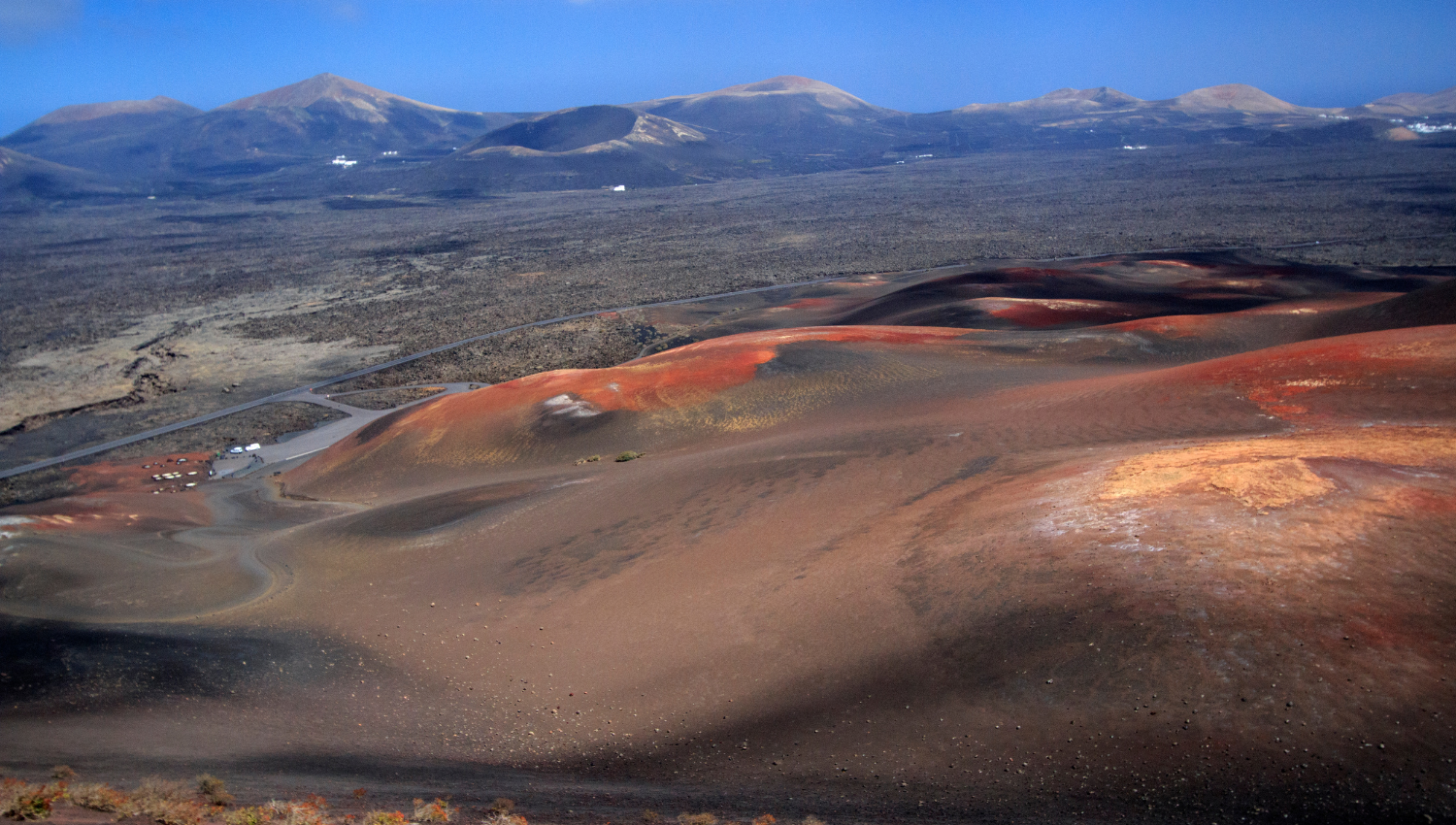 Vista panorámica de un paisaje volcánico con colinas de ceniza roja y negra, rodeadas por una extensa llanura de lava y montañas al fondo bajo un cielo despejado.