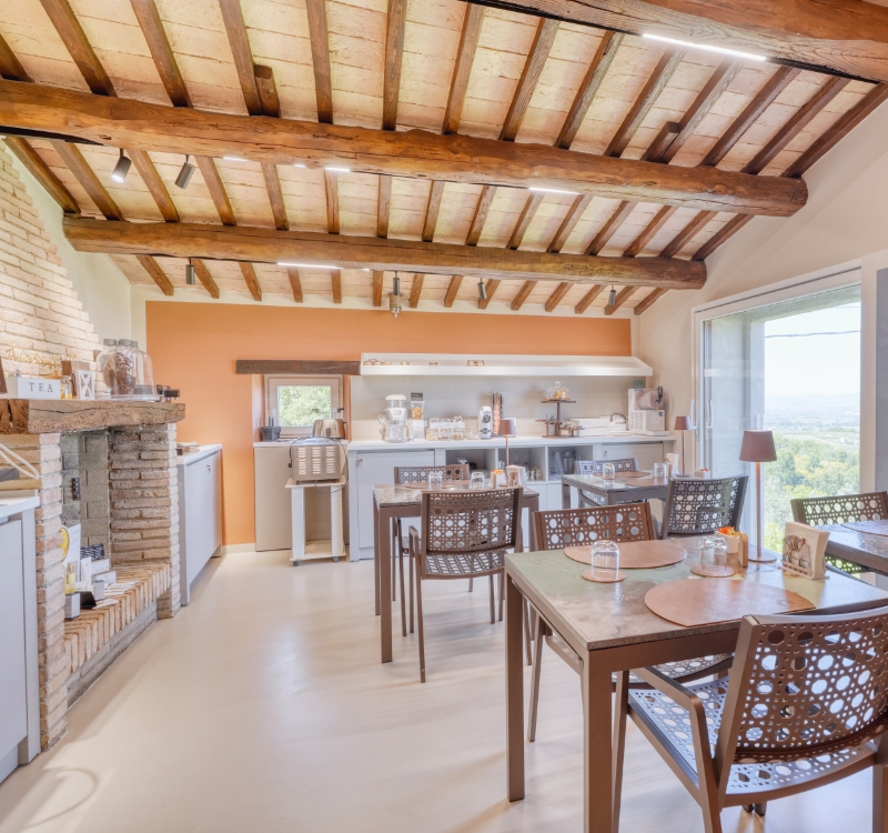 Sala colazione con travi a vista e camino caratteristico, dotata di quattro tavoli, area caffetteria, area buffet e ampia finestra con vista sull’uliveto. 