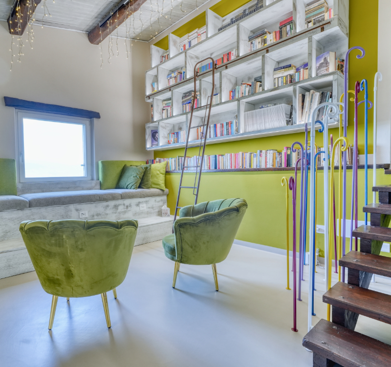Biblioteca con scaffali colmi di libri, arredata con due poltrone e panca con cuscini. Questa stanza è caratterizzata da una finestra con vista sull’uliveto e dettagli di design per il relax. 