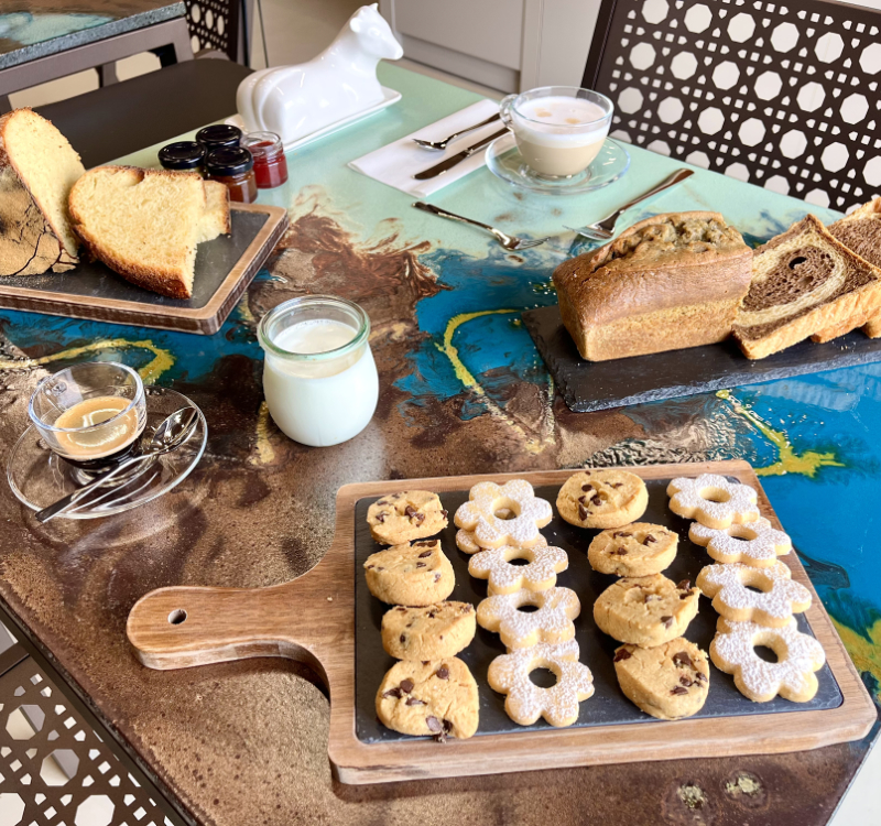 Tavolo della colazione con latte vegetale, espresso e cappuccino, accompagnati da pane fatto in casa, biscotti artigianali al burro d’arachidi con gocce di cioccolato e biscotti “Margheritine”, composta di cipolle, portaburro a forma di mucca e cake bicolore. 