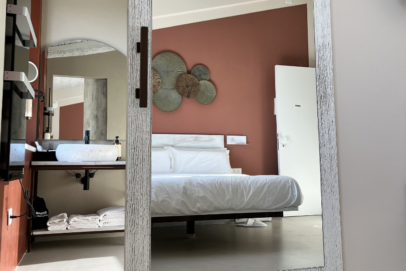 Camera Leccino, porta scorrevole del bagno con ampio specchio che riflette il letto sospeso. Nel bagno si intravede il lavabo, asciugamani e un’altro specchio. 