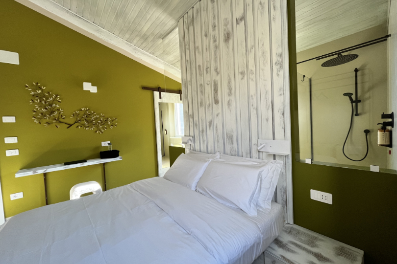 Camera Pendolino con letto sospeso, comodo pouf e scrivania e vetro attraverso il quale si intravede la doccia walk-in.