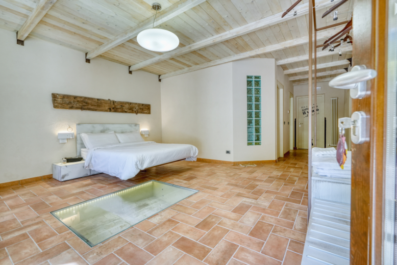 Camera Ascolano con letto matrimoniale sospeso, pavimento in resina e area vetrata che valorizza le antiche fondamenta del casale. 