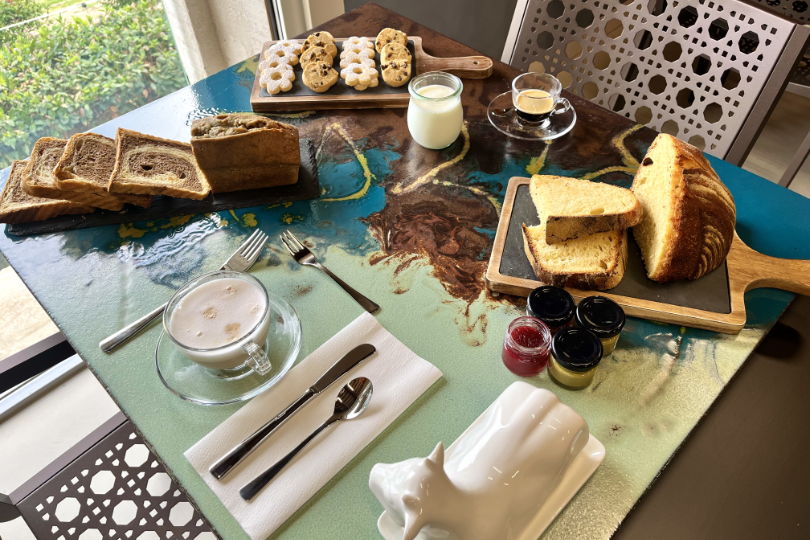 Tavolo della colazione con latte vegetale, espresso e cappuccino, accompagnati da pane fatto in casa, biscotti artigianali al burro d’arachidi con gocce di cioccolato e biscotti “Margheritine”, composta di cipolle, portaburro a forma di mucca e cake bicolore. 