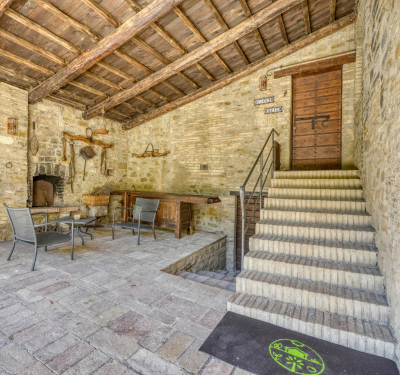 Portico con scala di accesso al piano superiore, attrezzato con posti a sedere dove potersi rilassare e tavolo originale. Barbecue e oggetti d’epoca appesi alle pareti. 